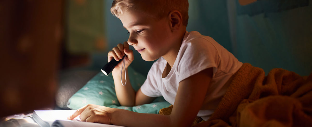 Un garçon lisant un livre avec une lampe torche à la main, allongé sur son lit avec une couverture